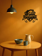 Decorative metal wall art with 'Welcome' on an orange wall above a wooden table with a bowl, mug, and pitcher.