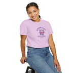 Person wearing a purple t-shirt with a graphic design and text, sitting on a stool against a white background.