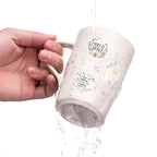 White mug with text and graphics being held by a hand, with water pouring out.